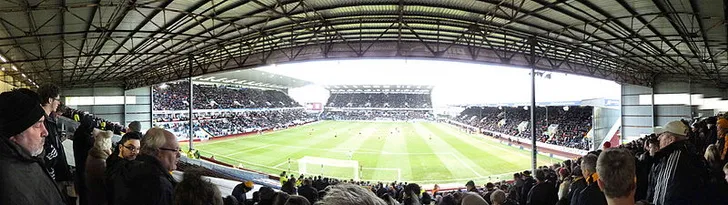 Toàn cảnh sân vận động Turf Moor nhìn từ khán đài, cho thấy bầu không khí cuồng nhiệt của một trận đấu bóng đá Anh, với các cổ động viên và kiến trúc đặc trưng của sân nhà Burnley.